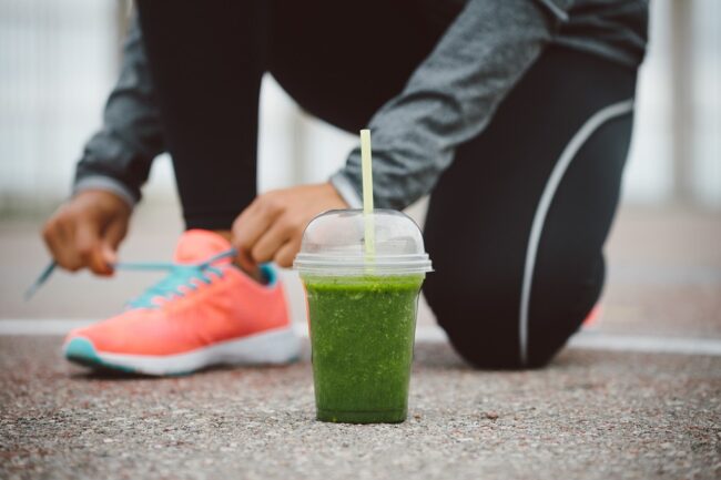 Detox smoothie drink and running footwear close up. City outdoor workout and fitness healthy nutrition concept. Female athlete tying sport shoes laces before training.
